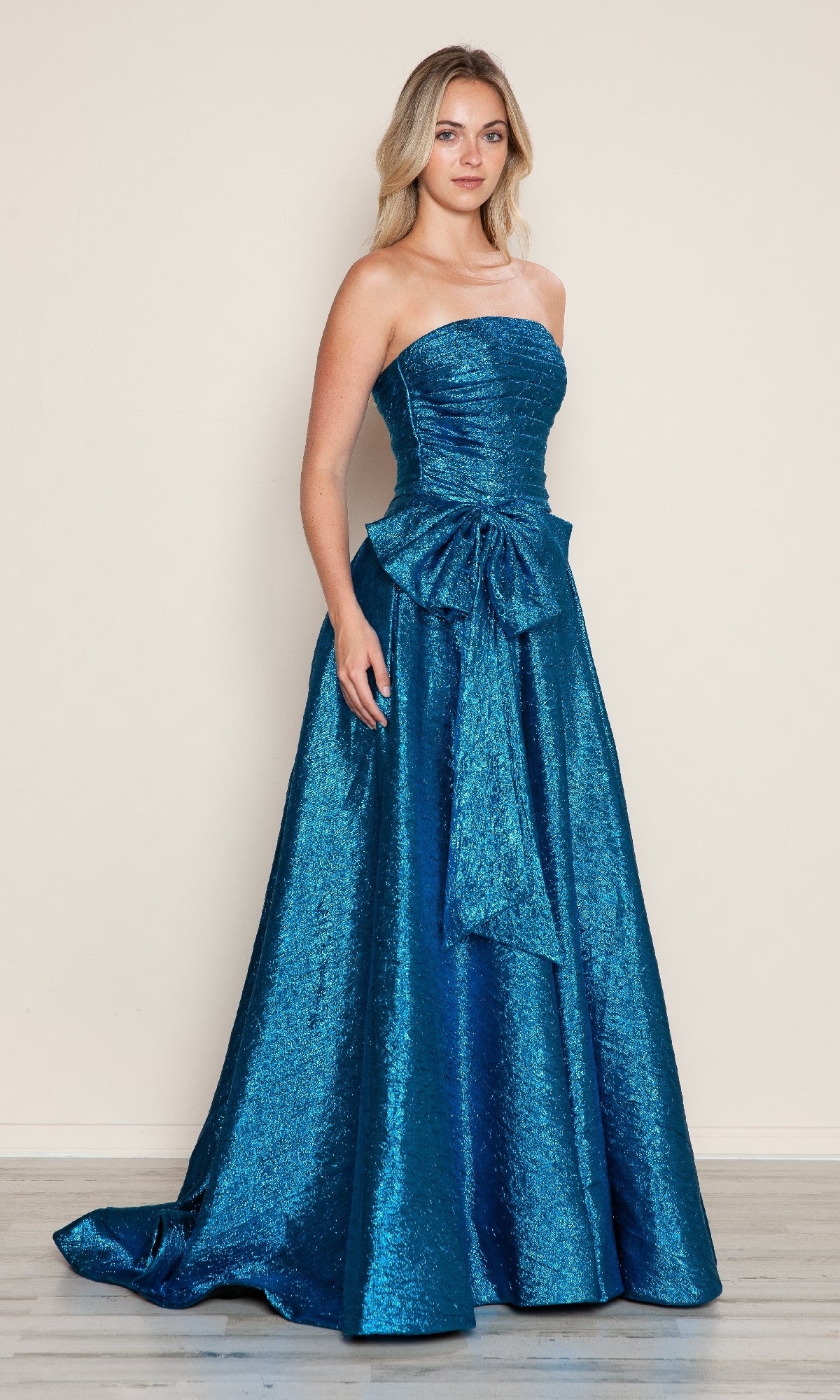 A woman models the Poly USA 9720 Side-Bow Strapless Prom Gown in metallic blue, showcasing its long glitter design and bold waist bow against a light, plain backdrop.